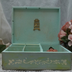 May include: A light blue wooden box with a decorative birdcage design on the lid. The box has six compartments and a white floral design on the bottom. The box is sitting on a white lace tablecloth.