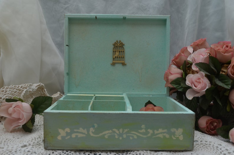 May include: A light blue wooden box with a decorative birdcage design on the lid. The box has six compartments and a white floral design on the bottom. The box is sitting on a white lace tablecloth.