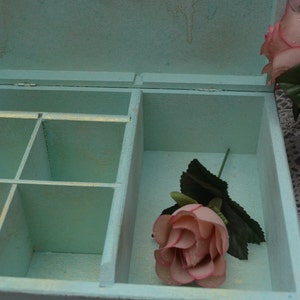 May include: A light blue wooden box with a hinged lid and compartments inside. The box is open and a single pink rose is inside. The box is sitting on a white lace surface.