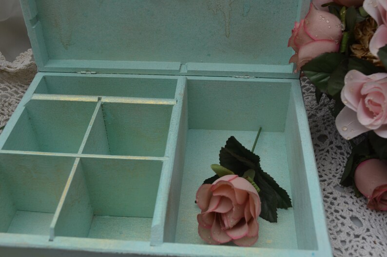 May include: A light blue wooden box with a hinged lid and compartments inside. The box is open and a single pink rose is inside. The box is sitting on a white lace surface.