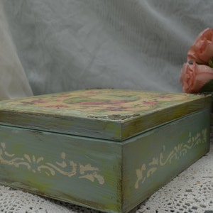 May include: A vintage-style wooden box with a light blue paint finish and a floral design on the lid. The box has a distressed look and is decorated with white painted details.