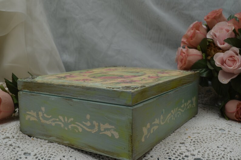May include: A vintage-style wooden box with a light blue paint finish and a floral design on the lid. The box has a distressed look and is decorated with white painted details.