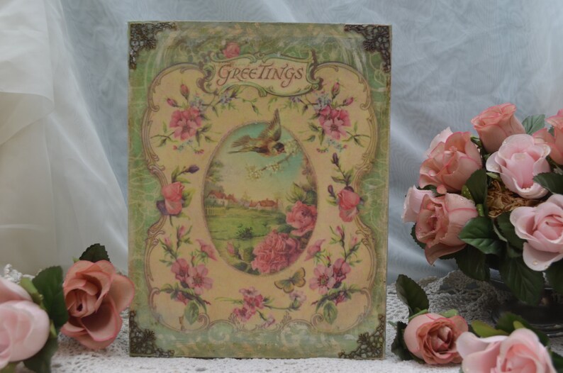May include: A vintage-style greeting card with a green and gold frame. The card features a floral design with a bird in flight and the word "Greetings" at the top.