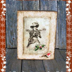 May include: A framed vintage image of a woman in a hat holding a bouquet of flowers. The frame is decorated with white lace and a small red bird is perched on the bottom.