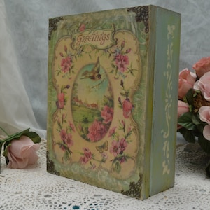 May include: A vintage-style wooden box with a floral design and the word "Greetings" on the front. The box is painted in shades of green and blue and features a bird and flowers.