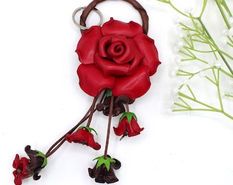 May include: A keychain featuring a large, sculpted red leather rose attached to a dark brown leather ring. Four smaller, dangling leather rosebuds in red and dark brown, each with green sepals, hang from thin leather cords attached to the main rose.