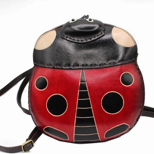 May include: A red leather ladybug-shaped crossbody bag with black spots and a black face. The bag has a brown adjustable strap.