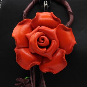 Puede incluir: Primer plano de una rosa de cuero naranja vibrante. La flor artesanal es el punto focal, con p&eacute;talos detallados y una apariencia realista. Est&aacute; unida a un fondo oscuro, posiblemente un bolso o accesorio.