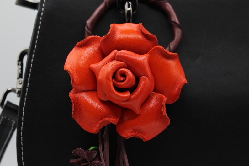 Puede incluir: Primer plano de una rosa de cuero naranja vibrante. La flor artesanal es el punto focal, con p&eacute;talos detallados y una apariencia realista. Est&aacute; unida a un fondo oscuro, posiblemente un bolso o accesorio.