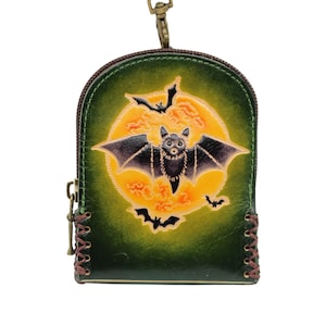 May include: A green and brown leather coin purse with a Halloween theme. The purse features a bat with outstretched wings in front of a yellow moon. Additional bats are flying around the moon. The purse has a zipper closure and a metal clasp.