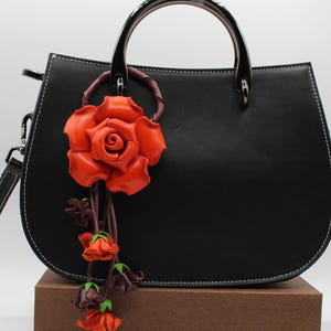 Puede incluir: Bolso de mano de cuero negro con forma curvada, costuras blancas y asa superior. Una gran rosa de cuero naranja con capullos m&aacute;s peque&ntilde;os cuelga del asa. El bolso tiene una correa de hombro desmontable.