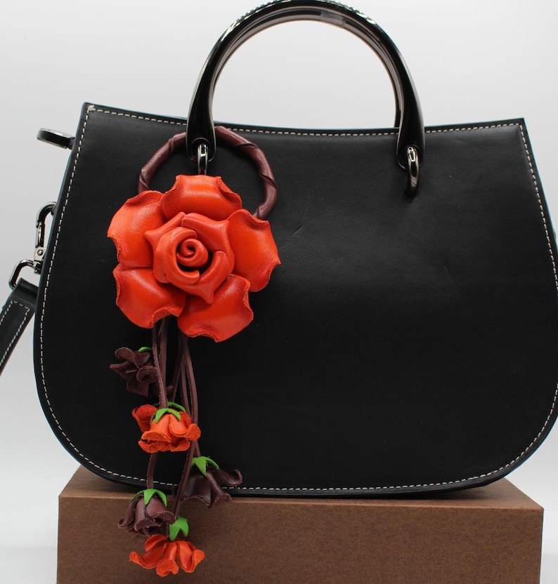Puede incluir: Bolso de mano de cuero negro con forma curvada, costuras blancas y asa superior. Una gran rosa de cuero naranja con capullos m&aacute;s peque&ntilde;os cuelga del asa. El bolso tiene una correa de hombro desmontable.