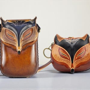 May include: Two brown leather bags, one a crossbody and the other a small purse, both featuring a fox face design. The fox faces are detailed with black and tan leather, with stitched accents. The crossbody bag has a long strap.