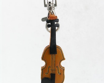 Puede incluir: Un llavero de cuero con forma de violín, que presenta un cuerpo de color tostado con detalles en negro en el mástil, el diapasón y el cordal. El instrumento está unido a un mosquetón giratorio de metal plateado y una anilla circular.