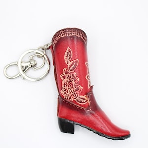 May include: Red leather cowboy boot keychain with floral detailing. The boot has a black heel and a silver-colored keyring and clasp. The boot is approximately 3 inches tall.