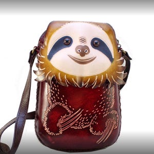 May include: A handcrafted leather crossbody bag featuring a sloth design. The bag has a smiling sloth face on the front with blue eyes and a light brown face. The body of the bag is a deep red color with etched details and a brown adjustable strap.