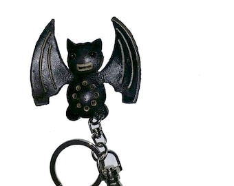 May include: A black leather bat keychain with a silver keyring and clasp. The bat has outstretched wings and a smiling face. The bat's body has several small, circular cutouts. A fun accessory.