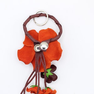 Puede incluir: Un llavero de cuero artesanal con un dise&ntilde;o de flor naranja. La flor tiene un tallo y detalles en marr&oacute;n oscuro, con un anillo y un cierre de bot&oacute;n de metal plateado. Peque&ntilde;os detalles de flores naranjas cuelgan de la parte inferior.
