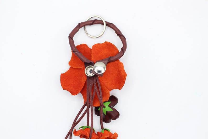 Puede incluir: Un llavero de cuero artesanal con un dise&ntilde;o de flor naranja. La flor tiene un tallo y detalles en marr&oacute;n oscuro, con un anillo y un cierre de bot&oacute;n de metal plateado. Peque&ntilde;os detalles de flores naranjas cuelgan de la parte inferior.