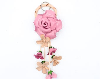 May include: A keychain featuring a large, layered pink leather rose attached to a braided beige leather ring with a metal split ring. Several thin beige leather cords hang from the rose, each ending in small, dangling pink and beige leather flowers.