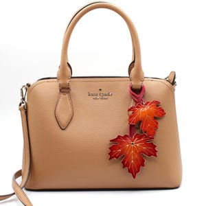 May include: Tan leather handbag with top handles and a detachable shoulder strap. The bag features a decorative charm of two orange and red maple leaves. The brand name "kate spade NEW YORK" is visible on the front.