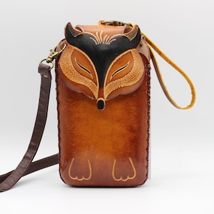 May include: Brown leather crossbody bag with a fox face design. The bag features a long adjustable strap and a small handle. The fox face is black and tan, with detailed stitching. The bag is approximately 7 inches tall.