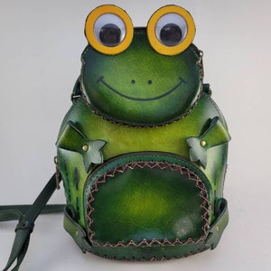 May include: Green leather frog backpack with yellow eyes and a smiling mouth. The backpack has a zippered pocket and a strap.
