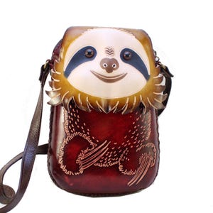 May include: A handcrafted leather crossbody bag featuring a smiling sloth face design. The bag has a brown leather strap and a burgundy body with etched details. The sloth's face is cream, brown, and black.