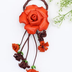 Puede incluir: Un accesorio floral para el cabello hecho a mano en cuero. El punto focal es una gran rosa naranja vibrante, complementada con flores m&aacute;s peque&ntilde;as de color naranja y marr&oacute;n oscuro. El dise&ntilde;o incluye tallos marrones y una banda circular de cuero marr&oacute;n.