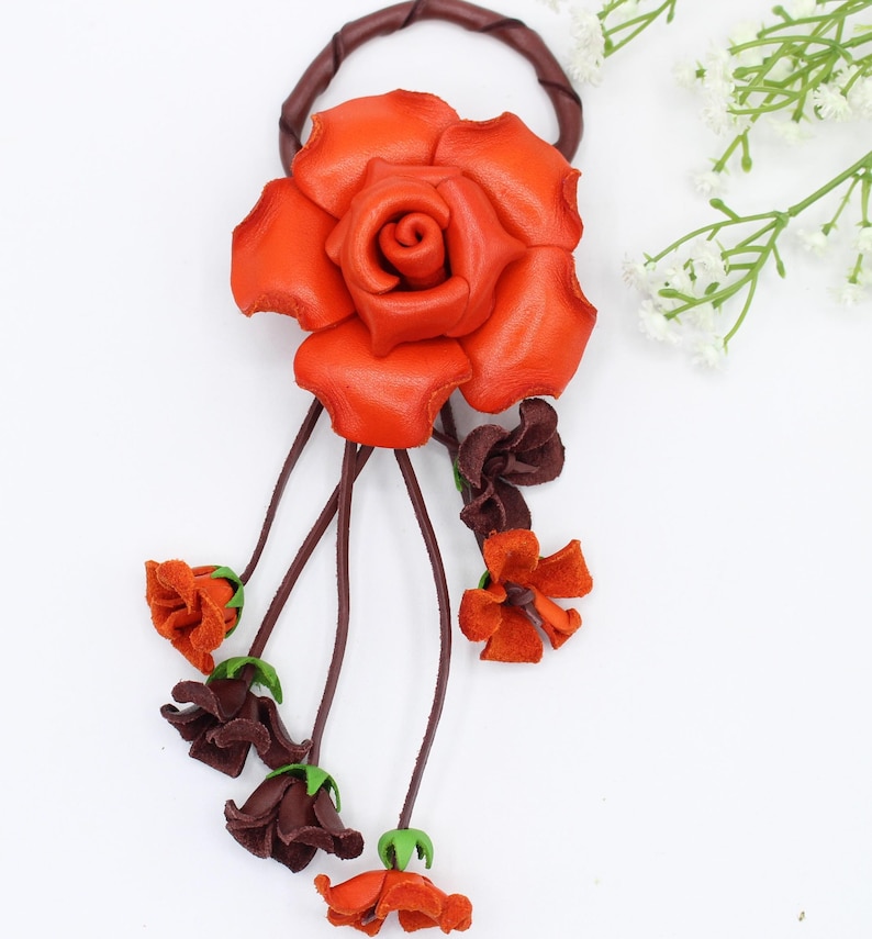 Puede incluir: Un accesorio floral para el cabello hecho a mano en cuero. El punto focal es una gran rosa naranja vibrante, complementada con flores m&aacute;s peque&ntilde;as de color naranja y marr&oacute;n oscuro. El dise&ntilde;o incluye tallos marrones y una banda circular de cuero marr&oacute;n.