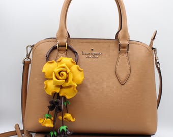 May include: A tan leather handbag with two top handles and a detachable shoulder strap. The bag features the text kate spade NEW YORK in gold lettering and a decorative charm consisting of a large yellow rose and smaller hanging flower buds.