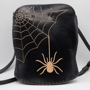 May include: Black crossbody bag featuring a spider web and spider design. The bag has a long, adjustable strap and a zipper closure. The spider web and spider are tan-colored, contrasting against the black leather.