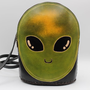May include: A green and black leather crossbody bag featuring an alien face design. The alien face has large black eyes, a small smile, and a yellow-green gradient. The bag has a black strap and a zipper closure.