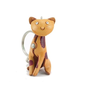 May include: A tan leather cat keychain with brown spots. The cat is sitting with its tail curled around its legs. It has a smiling face with white eyes and a black nose.