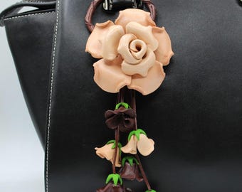 May include: A leather rose handbag charm in shades of tan and brown, with smaller flower accents in brown and green. The charm is attached to a black handbag, creating a stylish accessory.