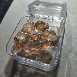 May include: A collection of seven raw, unpolished gemstones with translucent, amber-toned, and reddish-brown hues contained within a small, square, clear plastic hinged display box with a transparent lid.