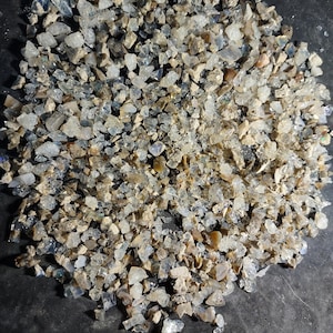 May include: A pile of small, irregularly shaped, translucent gemstones. The stones have a mix of colors, including clear, light blue, and tan. The gemstones are scattered across a dark surface, creating a textured appearance.