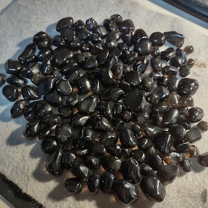 May include: A pile of polished, dark brown gemstones with a glossy finish. The stones vary in size and shape, and are scattered across a light gray surface. The gemstones appear to be smooth and rounded.