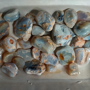 May include: A collection of smooth, tumbled blue chalcedony stones submerged in water. The stones exhibit translucent light blue hues with patches of opaque white and tan mineral inclusions, varying in size and irregular, rounded shapes.