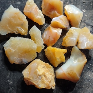 May include: A collection of raw, uncut opal gemstones. The stones vary in size and shape, displaying a range of colors from pale yellow to creamy white and orange. The opals have a rough, natural texture and are arranged on a dark surface.