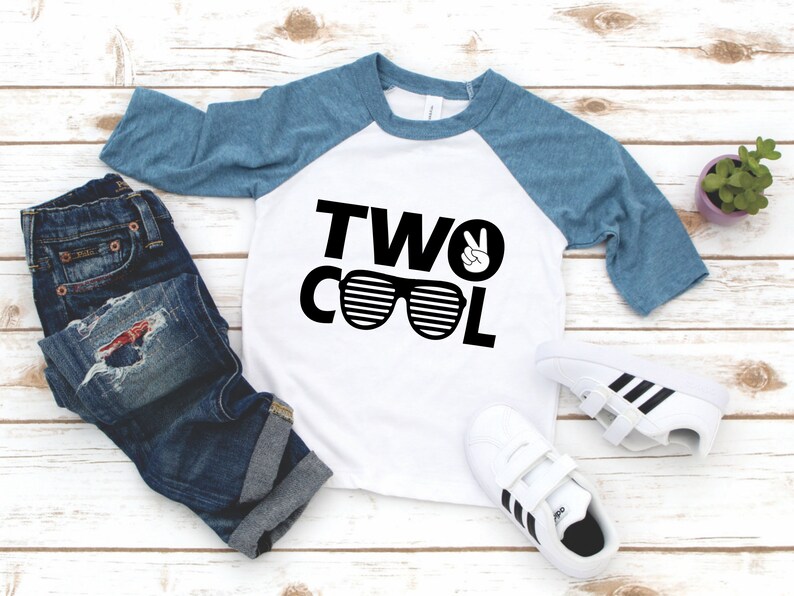 Może przedstawiać: Biało-niebieska koszulka bejsbolowa z napisem "TWO COOL" w kolorze czarnym, z symbolem pokoju i grafiką okular&oacute;w przeciwsłonecznych. Koszulka leży na białym drewnianym tle, obok pary białych but&oacute;w sportowych z czarnymi paskami i małą doniczką z rośliną.