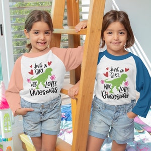 Może przedstawiać: Dw&oacute;jka dzieci w białych koszulkach z zielonym dinozaurem i napisem "Just a Girl Who Loves Dinosaurs". Jedno dziecko ma na sobie r&oacute;żowo-białą koszulkę, a drugie niebiesko-białą.