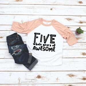 May include: A white and peach coloured raglan t-shirt with black text that reads "FIVE whole years of AWESOME".