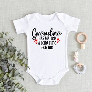 May include: White baby onesie with black text that reads "Grandma has waited a long time for me". There are four red hearts around the text.