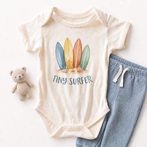 May include: Cream-colored baby onesie with a watercolor design of four surfboards in blue, yellow, orange, and green. The text "TINY SURFER" is below the surfboards. A pair of blue pants and a small teddy bear are next to the onesie.