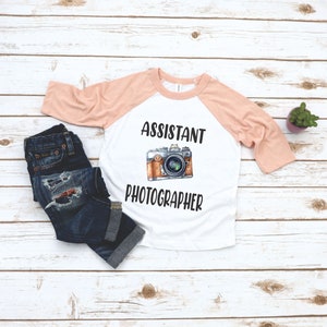 May include: A white and peach colored raglan t-shirt with the text "Assistant Photographer" and a cartoon camera graphic.