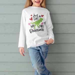 Może przedstawiać: Biała koszula z długim rękawem z zielonym dinozaurem i napisem "Just a girl who loves dinosaurs". Koszula ma r&oacute;żowe kwiaty i czerwone serca.