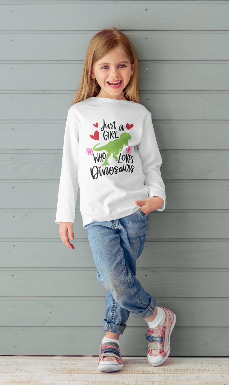 Może przedstawiać: Biała koszula z długim rękawem z zielonym dinozaurem i napisem "Just a girl who loves dinosaurs". Koszula ma r&oacute;żowe kwiaty i czerwone serca.
