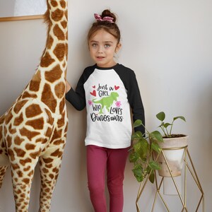 Może przedstawiać: Mała dziewczynka w biało-czarnej koszulce bejsbolowej z napisem "Just a girl who loves dinosaurs" i zielonym dinozaurem. Ma na sobie r&oacute;żowe legginsy i stoi obok pluszowej żyrafy.