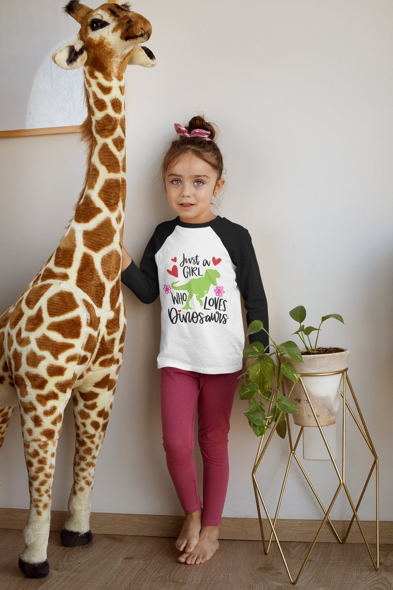 Może przedstawiać: Mała dziewczynka w biało-czarnej koszulce bejsbolowej z napisem "Just a girl who loves dinosaurs" i zielonym dinozaurem. Ma na sobie r&oacute;żowe legginsy i stoi obok pluszowej żyrafy.